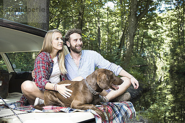 Lächelndes junges Paar mit Hund sitzt im Auto an einem Bach im Wald
