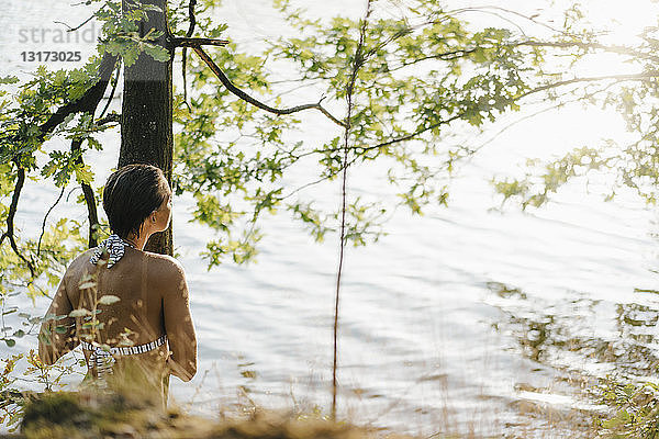 Rückansicht einer Frau im Bikini am Baumstamm an einem See