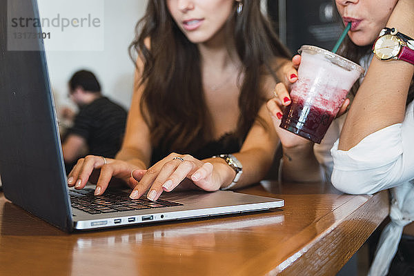 Junge Frau trinkt Smoothie in einem Café  während ihre Freundin einen Laptop benutzt  Teilansicht