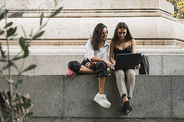 Zwei Freunde sitzen mit einem Laptop nebeneinander an einer Wand