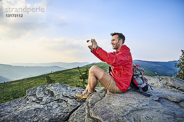 Mann sitzt auf einem Felsen und fotografiert mit seinem Handy während einer Wanderung in den Bergen