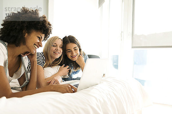 Drei glückliche Freundinnen liegen im Bett und teilen sich einen Laptop