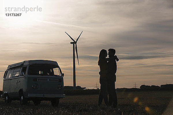 Junges Paar küsst sich in der Abenddämmerung vor einem Wohnmobil mit Windrad im Hintergrund