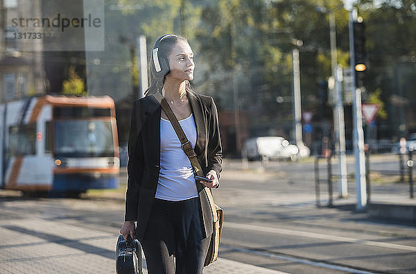 Junge Frau mit Kopfhörern und Handy an der Straßenbahnhaltestelle
