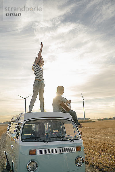 Glückliches Paar mit Gitarre auf dem Dach eines Wohnmobils in ländlicher Landschaft