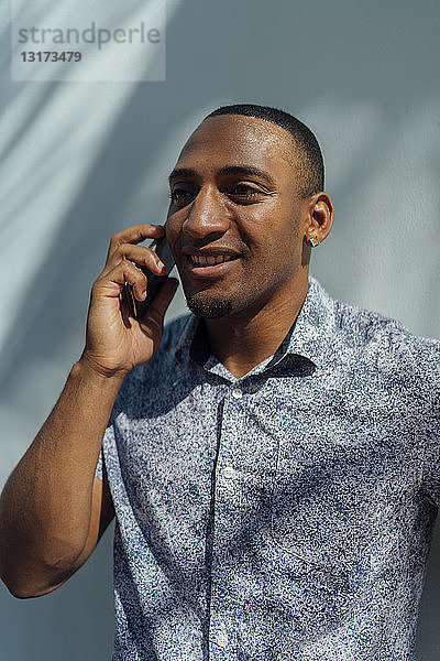 Lächelnder junger Mann im Hemd  der mit seinem Handy an einer Wand telefoniert
