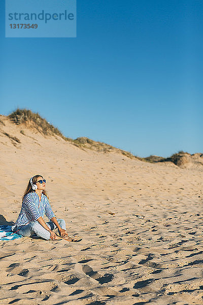 Portugal  Aveiro  Frau sitzt in der Nähe einer Stranddüne und hört Musik mit Kopfhörern