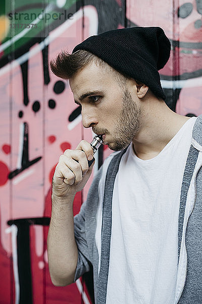 Junger Mann steht vor Graffiti und raucht elektronische Zigarette