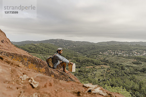 Junger Mann mit Rucksack  sitzt auf einem Berg und schaut auf die Aussicht