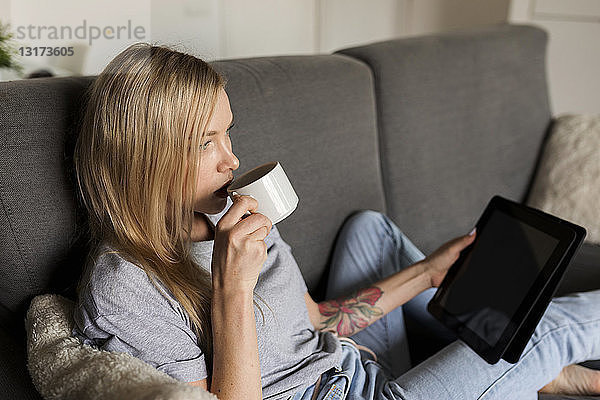 Junge Frau sitzt auf Couch  trinkt Kaffee und hält Tablette