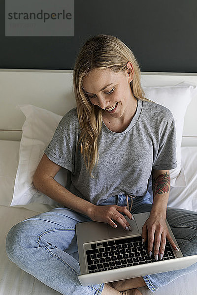 Glückliche junge Frau sitzt mit Laptop im Bett