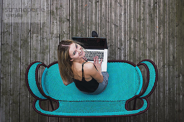 Lächelnde junge Frau sitzt auf türkisfarbener Couch auf der Terrasse mit Laptop  Draufsicht