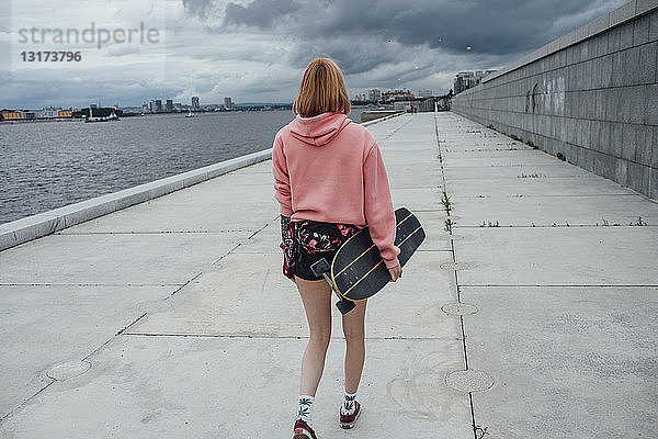 Rückansicht einer jungen Frau  die ein Carver-Skateboard hält und am Flussufer spazieren geht