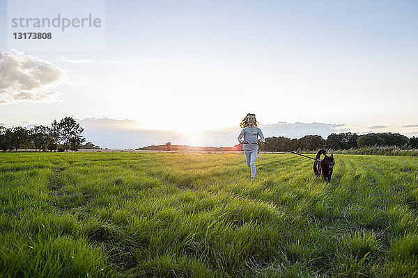 Mädchen mit einem Hund  das bei Sonnenuntergang über ein Feld läuft