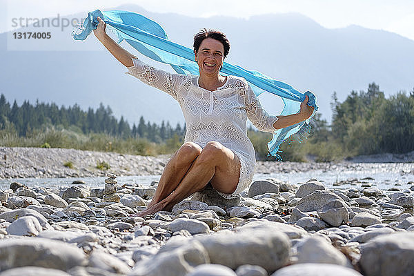 Glückliche Frau hält schwingenden blauen Schal am Flussufer  Oberbayern  Deutschland