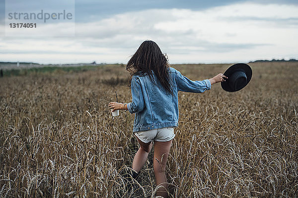 Rückenansicht einer jungen Frau mit Getränk und Hut beim Spaziergang im Maisfeld