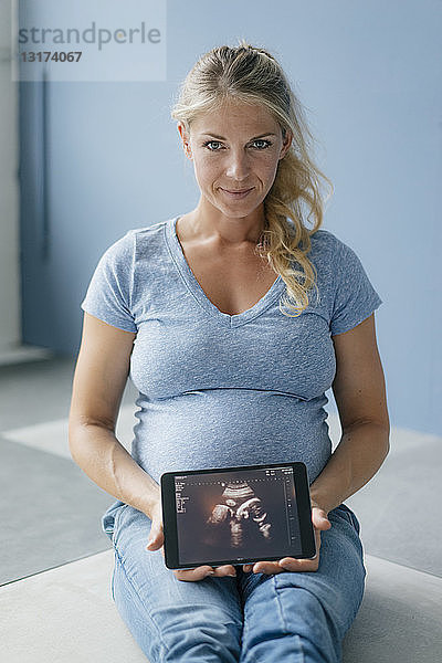 Porträt einer lächelnden schwangeren Frau  die auf dem Boden sitzt und ein Ultraschallbild auf einem Tablett zeigt