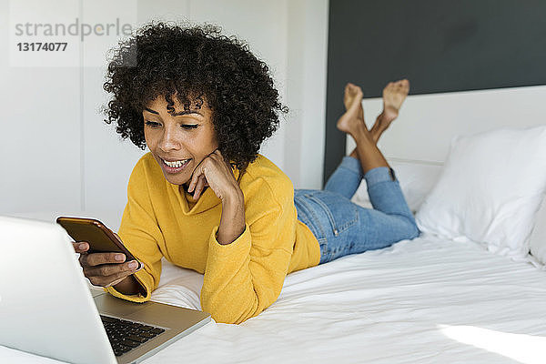Lächelnde Frau liegt im Bett und benutzt Handy und Laptop