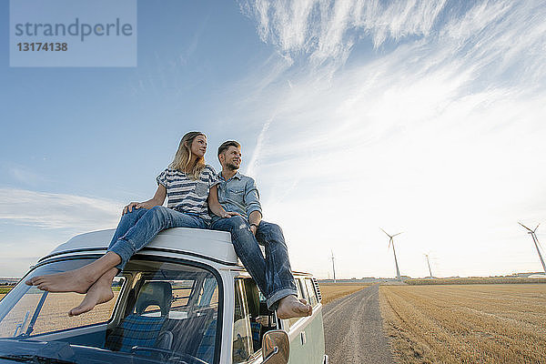 Auf Wohnmobil sitzendes Ehepaar in ländlicher Landschaft mit Windturbinen im Hintergrund