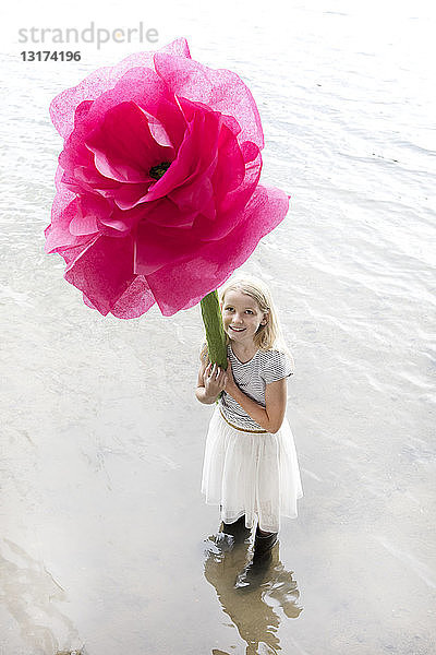 Porträt eines lächelnden blonden Mädchens  das in einem See steht und eine überdimensionale rosa Kunstblume hält