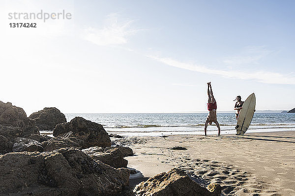 Frankreich  Bretagne  junger Mann macht einen Handstand und Frau hält ein Surfbrett am Strand