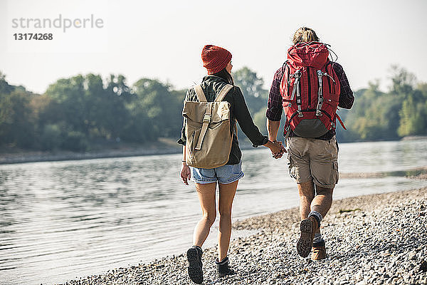 Rückansicht eines jungen Paares mit Rucksäcken  das Hand in Hand am Flussufer geht