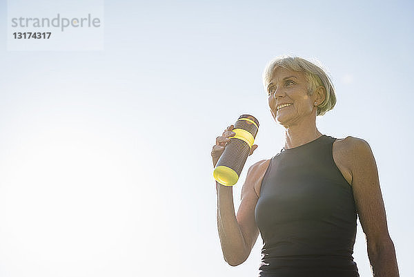 Lächelnde sportliche ältere Frau hält Flasche unter blauem Himmel