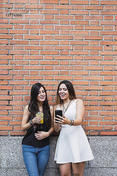 Mädchen-Freunde stehen vor einer Backsteinmauer und schauen auf ein Smartphone