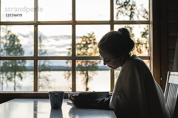 Finnland  Lappland  junge Frau sitzt am Fenster an einem See und schaut in ihr Tagebuch