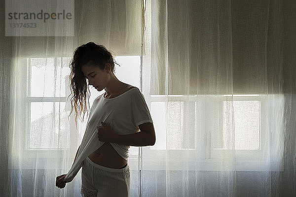 Junge Frau steht in weißer Unterwäsche vor dem Fenster