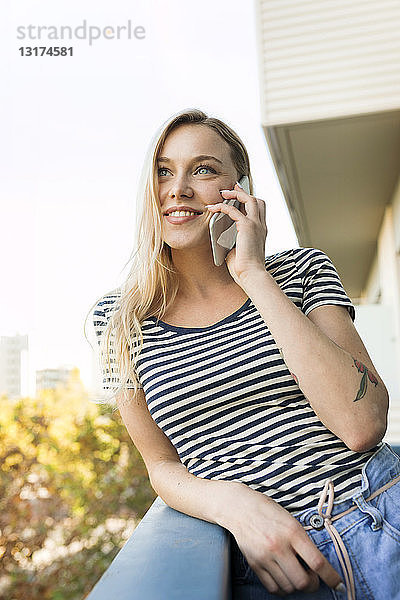 Lächelnde junge Frau telefoniert per Handy auf dem Balkon