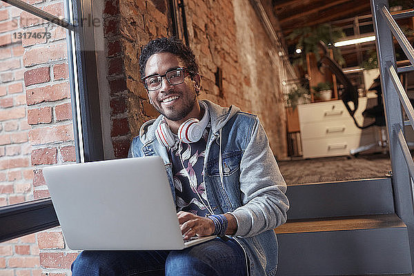 Junger Mann mit Kopfhörern sitzt auf der Bürotreppe und benutzt einen Laptop