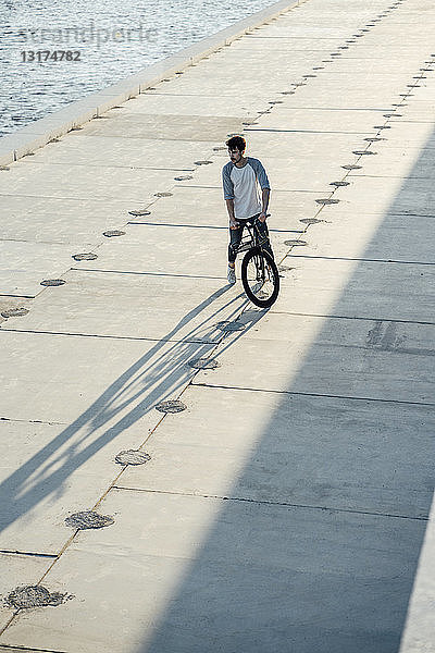 Junger Mann mit Pendler-Fixie-Fahrrad macht auf Uferpromenade am Fluss eine Pause