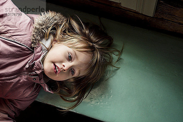 Porträt eines kleinen Mädchens  das auf einer Holzbank liegt und im Haus einen Anorak trägt