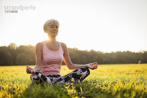 Ältere Frau in Lotus-Position auf ländlicher Wiese bei Sonnenuntergang