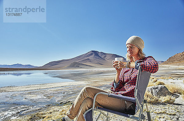 Bolivien  Laguna Colorada  Frau sitzt auf einem Campingstuhl am Seeufer und trinkt aus einer Tasse