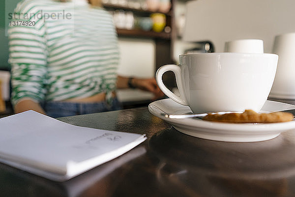 Tasse Kaffee und Notizblock auf Tisch in einem Café mit Frau im Hintergrund