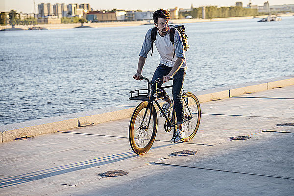 Junger Mann mit Rucksack auf dem Fahrrad auf der Uferpromenade am Flussufer