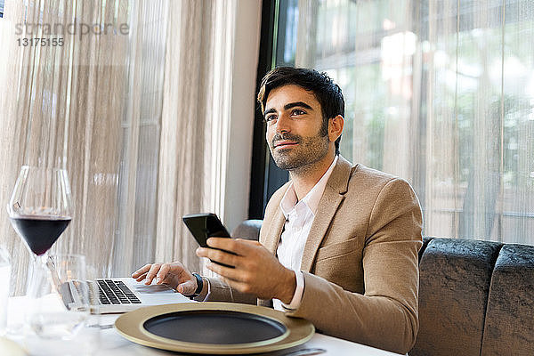 Lächelnder Mann sitzt am Tisch in einem Restaurant und benutzt Laptop und Handy