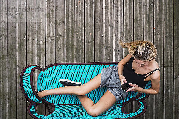 Junge Frau sitzt auf türkisfarbener Couch auf der Terrasse mit Smartphone  Draufsicht