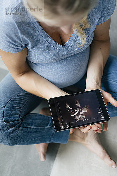 Schwangere Frau sitzt auf dem Boden und betrachtet Ultraschallbild auf Tablette
