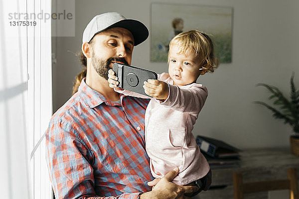Vater hält seine kleine Tochter auf dem Arm und spielt mit seinem Smartphone