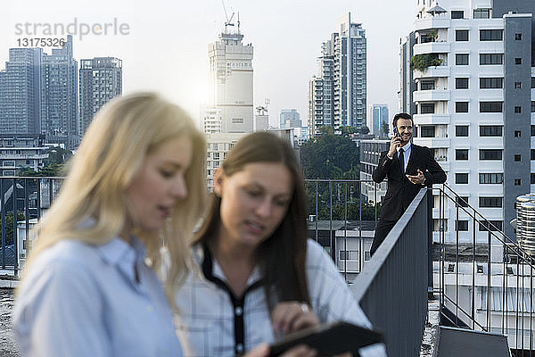 Geschäftsmann spricht auf Smartphone  während weibliche Kollegen auf dem Dach der Stadt auf das Tablet schauen