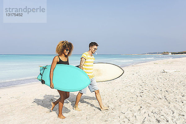 Paar  das am Strand spazieren geht und Surfbretter trägt