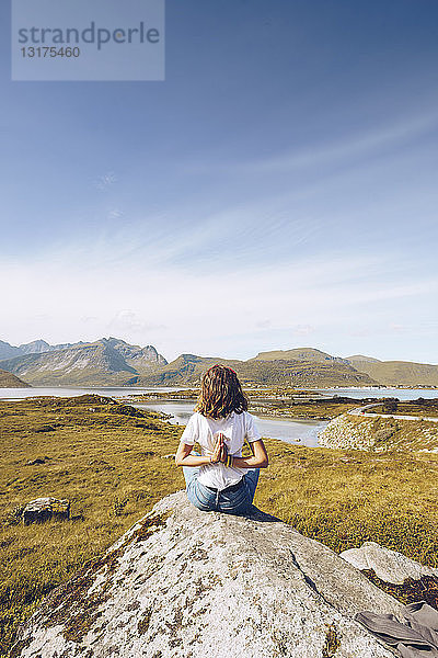 Norwegen  Lofoten  Rückenansicht einer jungen Frau  die auf einem Felsen sitzt und Yoga-Übungen macht
