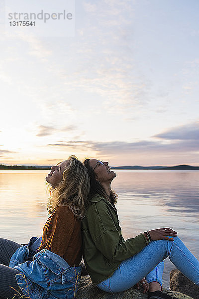 Finnland  Lappland  zwei glückliche junge Frauen sitzen Rücken an Rücken am Seeufer in der Dämmerung
