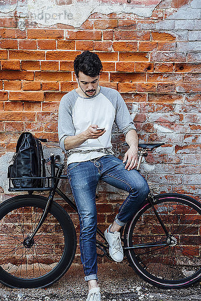 Junger Mann mit Pendler-Fixie-Fahrrad steht mit Handy und Kopfhörern an Ziegelmauer