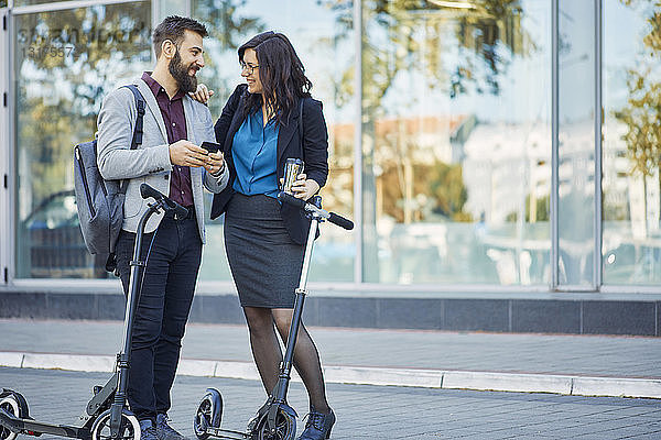 Lächelnder Geschäftsmann und Geschäftsfrau mit Rollern im Gespräch auf dem Bürgersteig