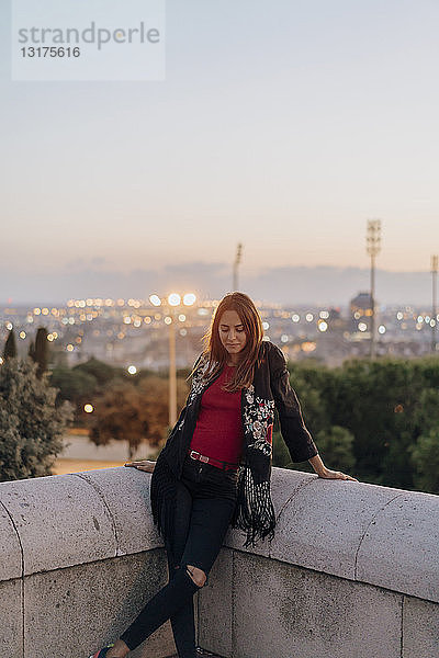 Spanien  Barcelona  Montjuic  junge Frau  die in der Abenddämmerung mit Stadtlichtern im Hintergrund an eine Wand gelehnt