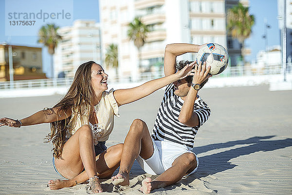 Glückliches junges Paar sitzt am Strand und spielt mit einem Ball